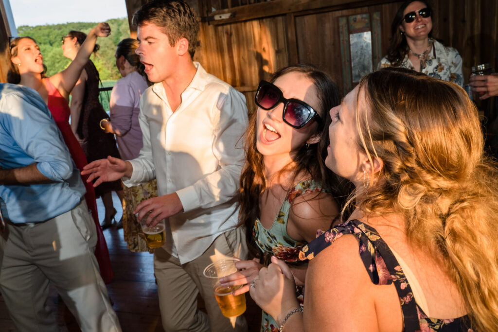 A group of people dance and sing at a lively indoor party. Two women in front smile, one wearing sunglasses. Others hold drinks, enjoying themselves with sunlight streaming in.