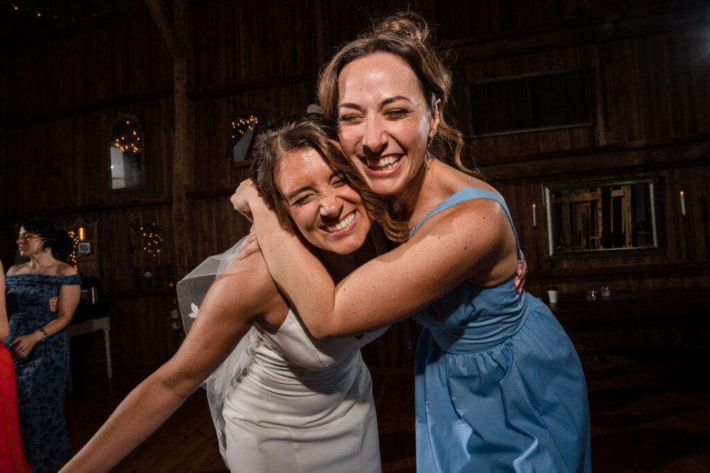 Two women, one in a white dress and veil and the other in a light blue dress, smile and hug closely at a wedding reception in a warmly lit wooden venue.