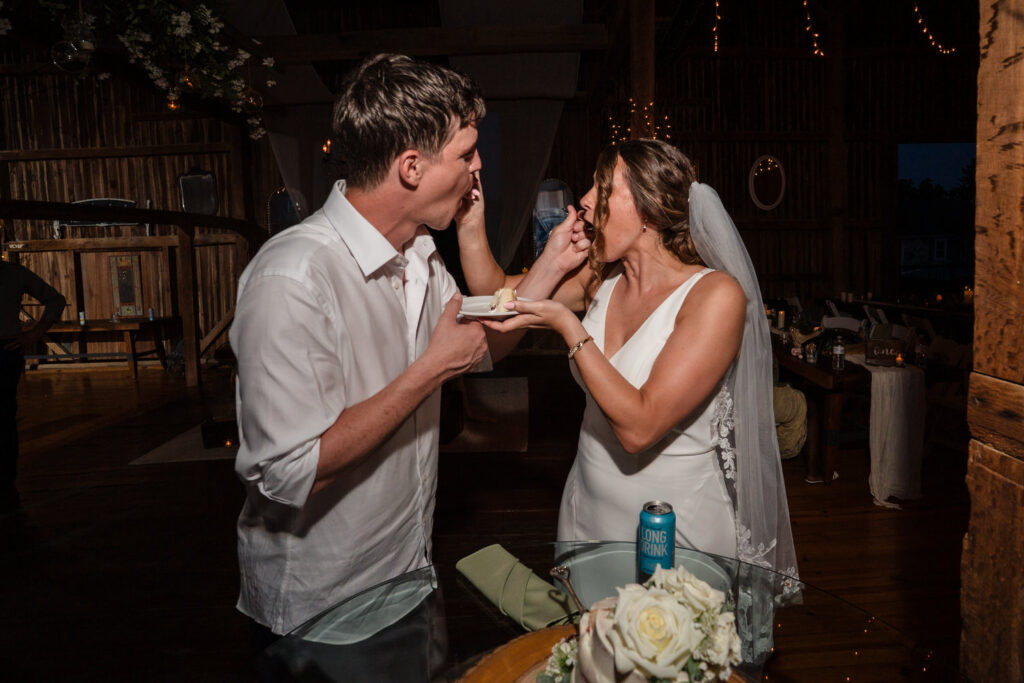 A bride and groom feed each other cake at their wedding reception, standing close and smiling, with a rustic wooden background and fairy lights.