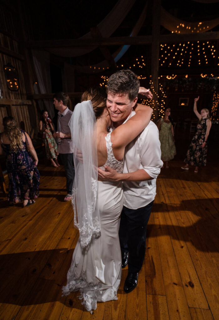 A bride in a white dress and veil hugs a man in a white shirt as they smile and dance on a wooden floor, surrounded by wedding guests under string lights.