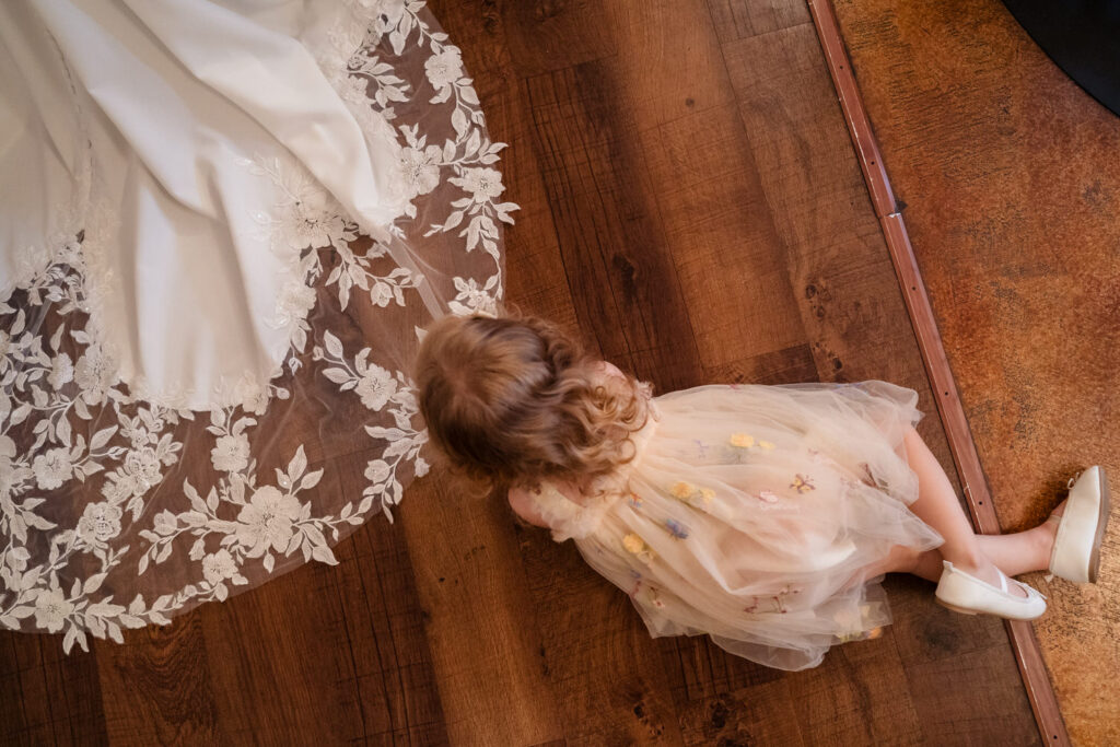 A young child in a light dress sits on a wooden floor, gazing at the lace train of a white wedding gown nearby.