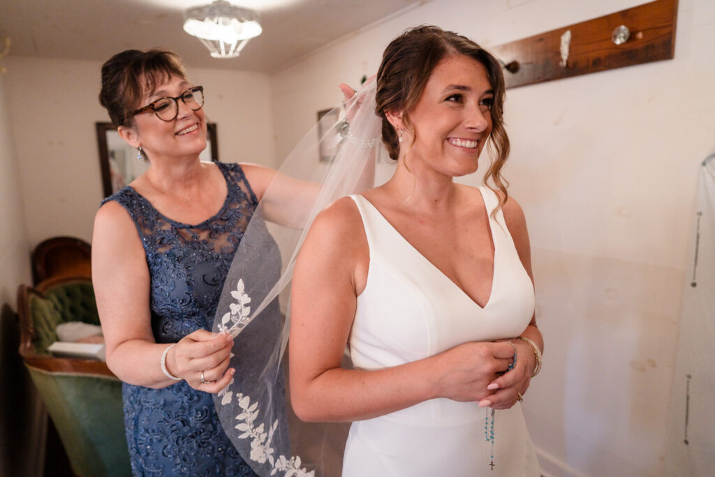 A woman in a blue lace dress helps a smiling bride in a white gown adjust her veil in a room, both looking happy and excited.
