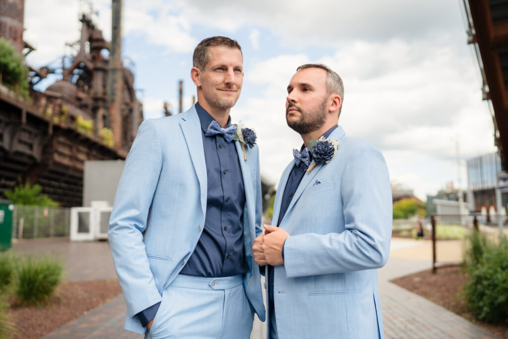 Two men in matching light blue suits with dark shirts and floral boutonnieres stand side by side outdoors, looking confident. An industrial structure and greenery are visible in the background under a cloudy sky.