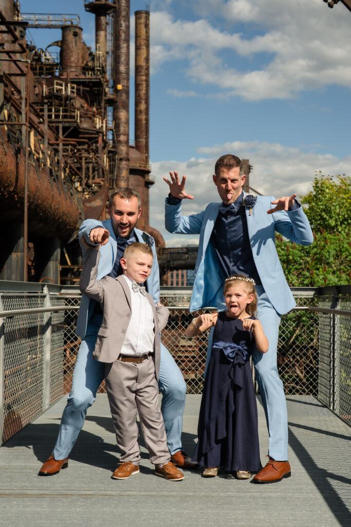 Two men in light blue suits and two children pose playfully on a walkway by an industrial backdrop, with the men making silly gestures and the kids smiling at the camera.