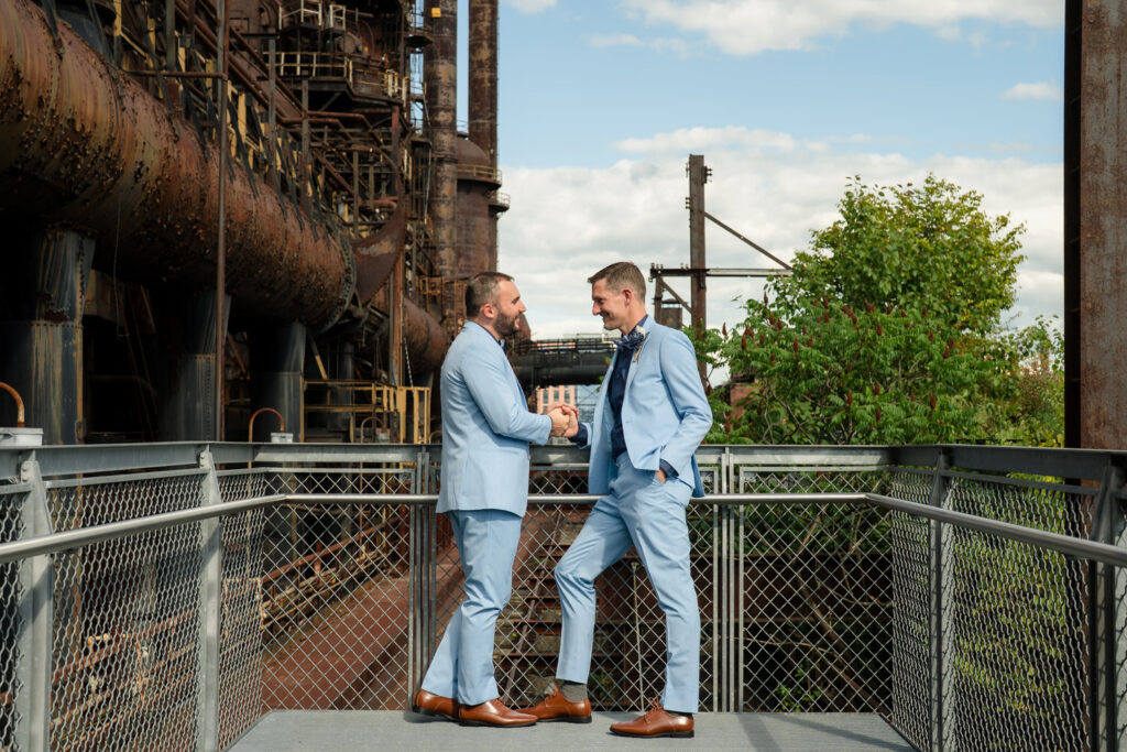Two men in light blue suits smile and shake hands on a metal platform, with rusted industrial structures and green trees in the background under a partly cloudy sky.