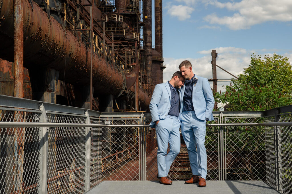 Two men in light blue suits stand close together on an outdoor metal platform, with one resting his forehead on the others. Behind them are rusted industrial structures and a blue sky with clouds.