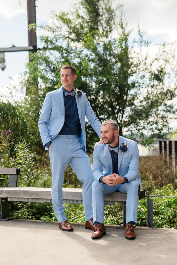 Two men in light blue suits, navy shirts, and brown shoes pose outdoors. One stands with a hand in his pocket, while the other sits on a bench. Both wear boutonnières and smile. Green trees and sky fill the background.