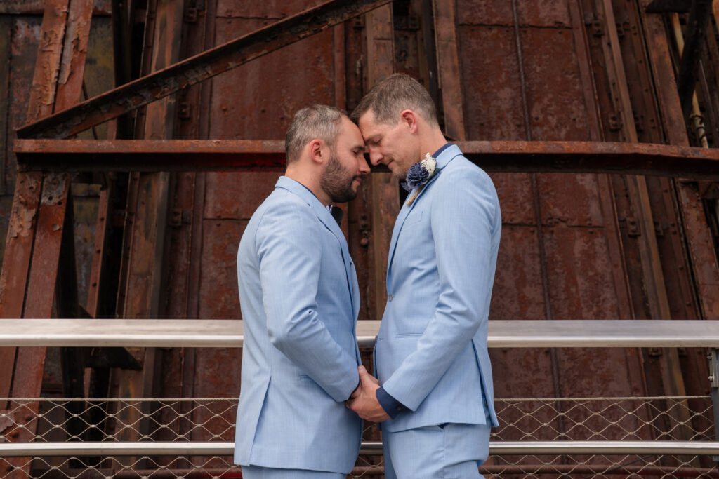 Two men in matching light blue suits stand face to face, holding hands and touching foreheads, against a rustic industrial background.