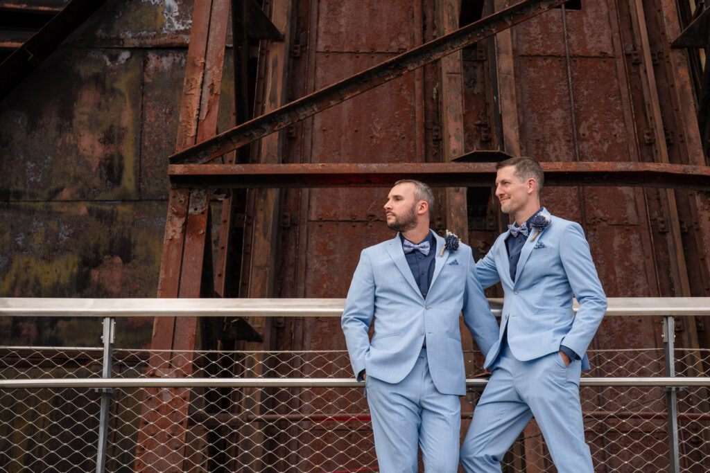 Two men in matching light blue suits stand side by side, posing confidently in front of a large, rusted industrial structure and metal railings.