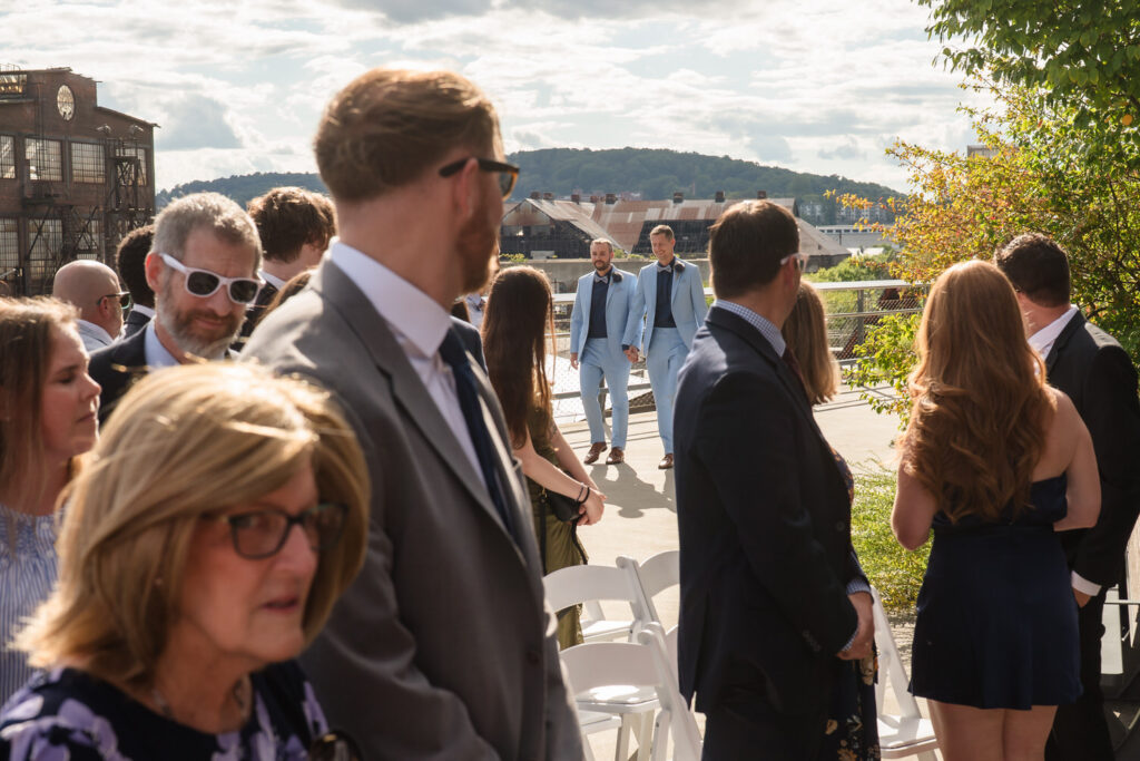 Wedding guests stand and turn as two grooms in light suits walk down an outdoor aisle toward them on a sunny day, with a scenic backdrop of hills and industrial buildings.