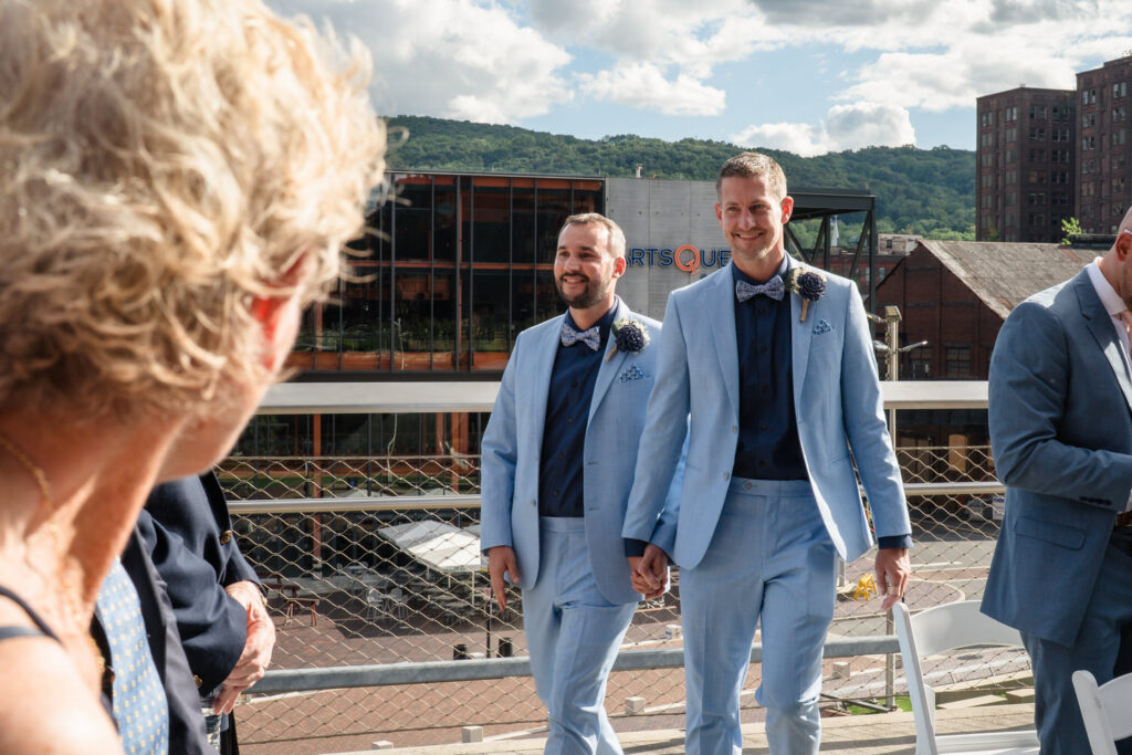 Two men in matching light blue suits walk hand in hand outside at a wedding. A guest in the foreground looks toward them. Buildings and a blue sky with clouds are in the background.