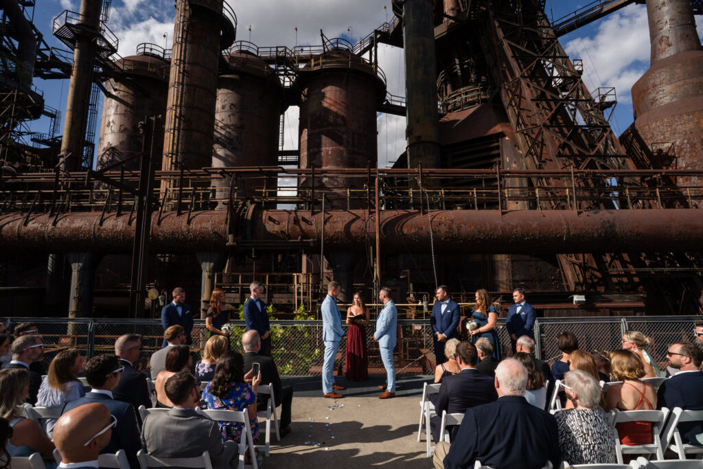 A wedding ceremony takes place outdoors in front of a large, rusted industrial structure, with guests seated and a couple standing at the altar under a sunny sky.