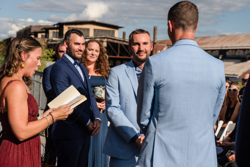 Two men in light blue suits hold hands at an outdoor wedding ceremony. A woman in a red dress reads from a book. Guests and members of the wedding party stand nearby, smiling and watching.