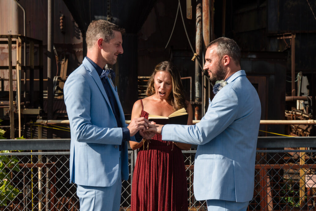 Two men in matching light blue suits exchange rings during a wedding ceremony, with an officiant in a maroon dress standing between them, reading from a book. The background features industrial metal structures.