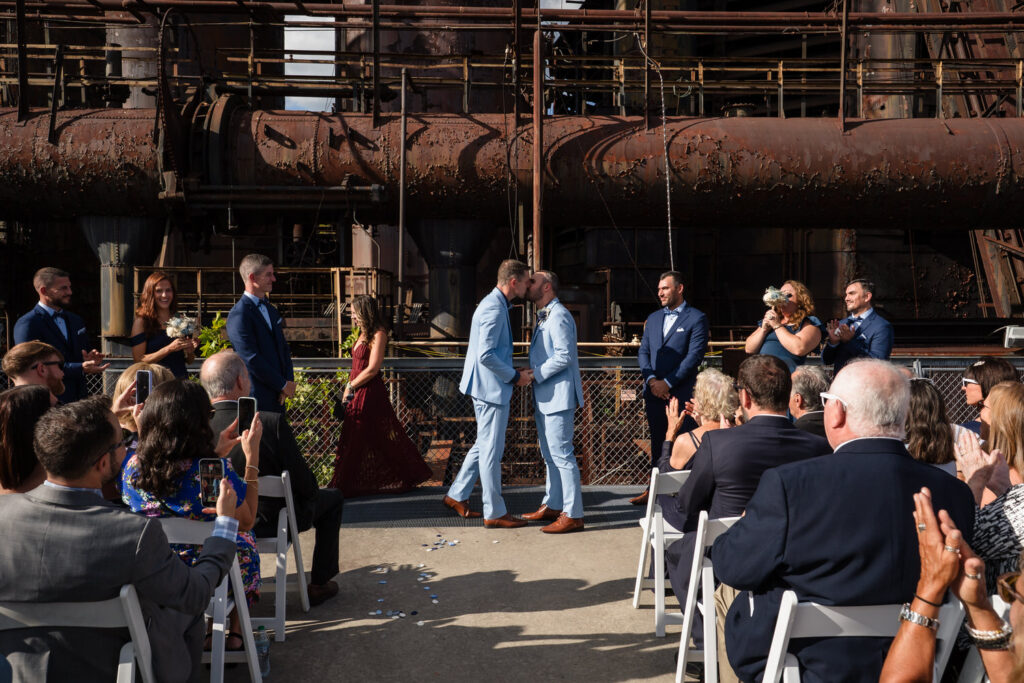 Two grooms in matching blue suits kiss at an outdoor wedding ceremony, surrounded by their wedding party and guests, with a rusted industrial structure in the background.