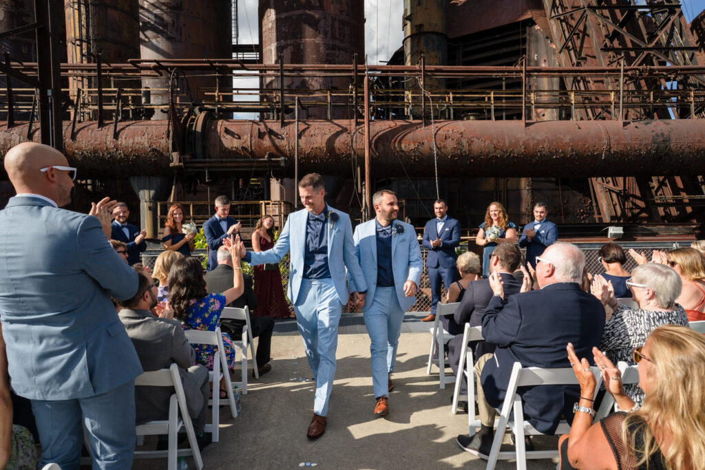 Two grooms in light blue suits walk hand in hand down the aisle, smiling as guests applaud at an outdoor wedding with an industrial, rusted metal backdrop.