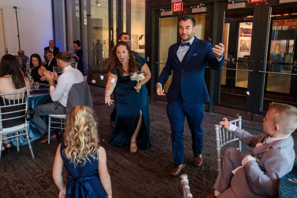 A woman and man in formal attire enter a room excitedly, reaching out to a young girl in a blue dress, while seated guests watch and smile.