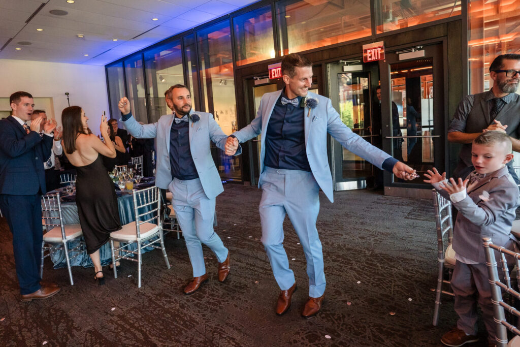 Two men in light blue suits joyfully enter a room holding hands, smiling as guests clap for them. A young boy in a matching suit stands by, reaching out for a high five. The event appears to be a celebration or wedding.