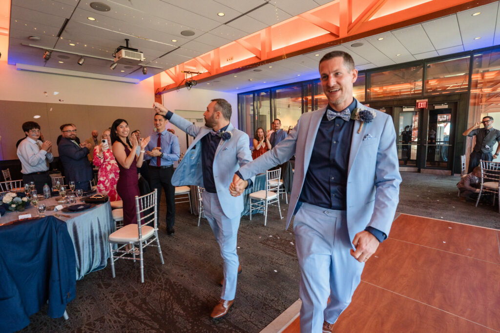 Two men in light blue suits, holding hands and smiling, enter a brightly lit room as guests stand and applaud at a celebratory event, possibly a wedding reception.