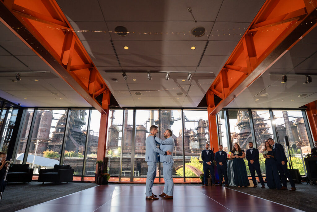 Two grooms share a first dance in a modern venue with large windows, revealing an industrial backdrop. Wedding party members stand nearby, watching and smiling.