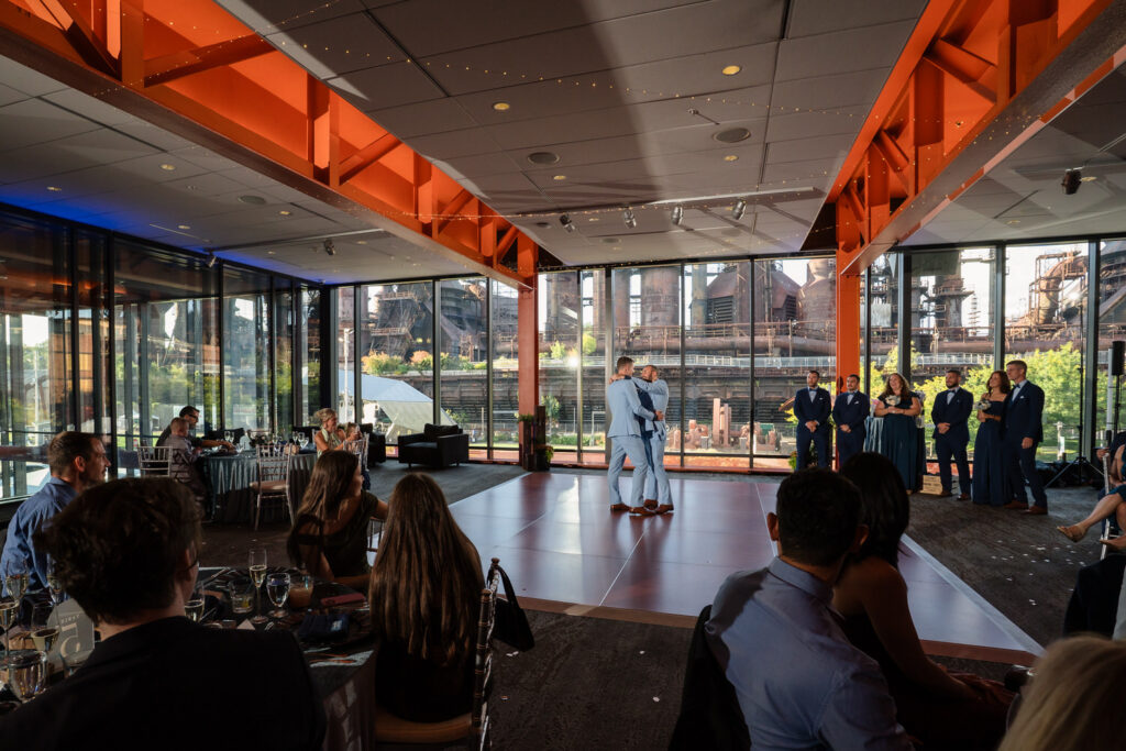 A couple shares their first dance in a modern venue with large windows, orange beams, and an industrial view outside; guests are seated and watching, while the wedding party stands nearby.