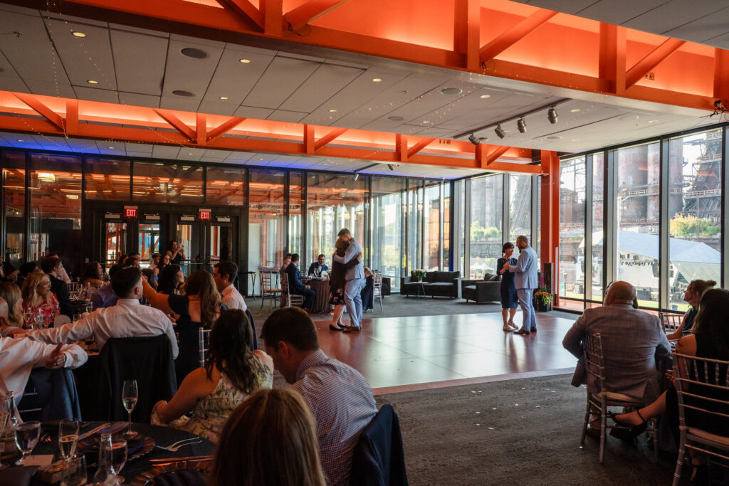A wedding reception with guests seated at tables watching couples dance on a spacious, sunlit floor surrounded by large windows and orange beams.