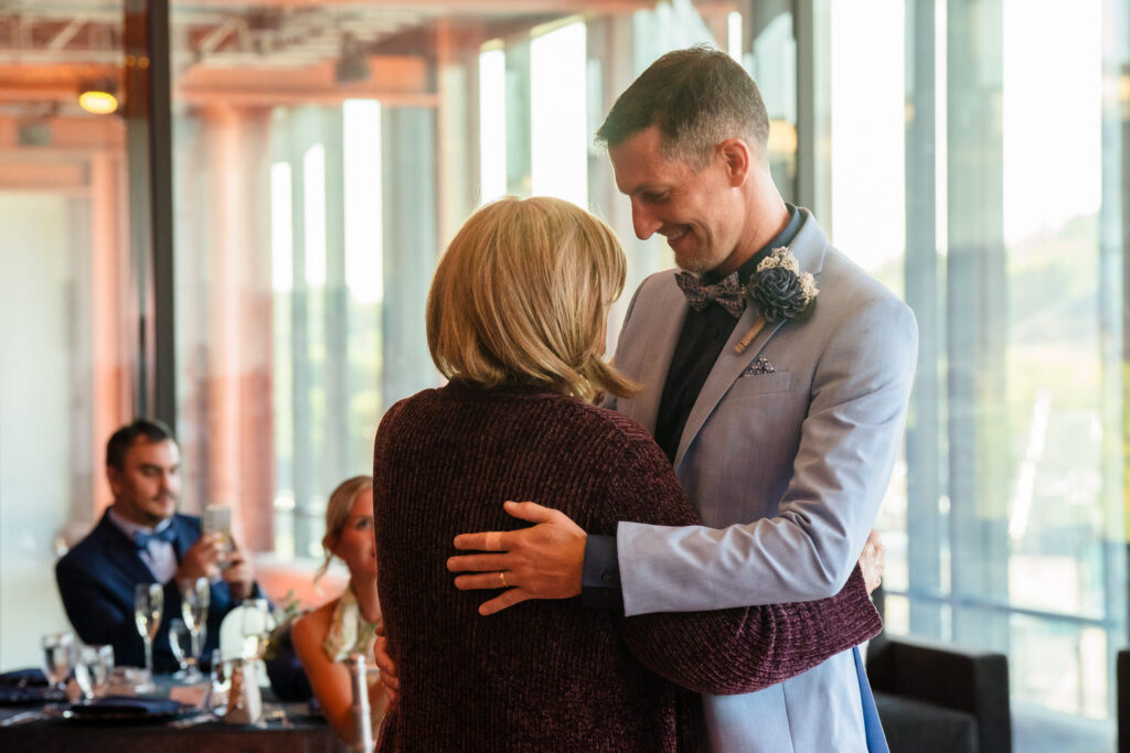 A man in a light blue suit and bow tie dances with a woman in a maroon sweater at a formal event, smiling at each other. Other guests sit at tables in the background, some watching and some talking.