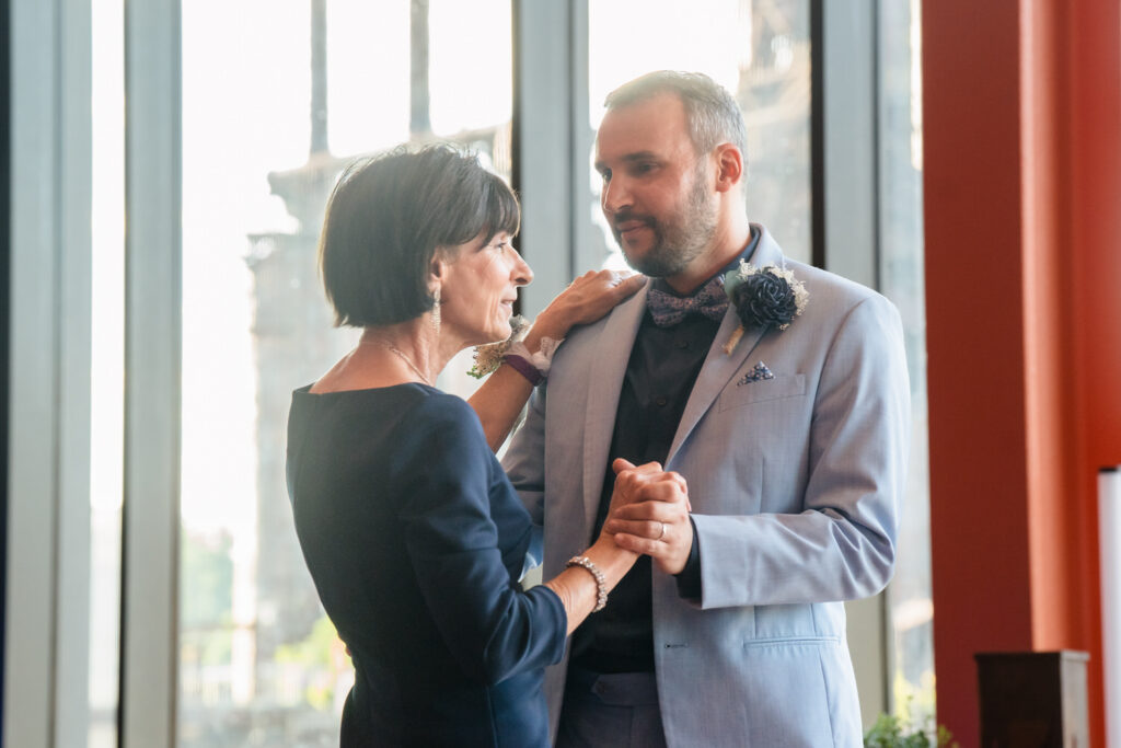A woman and a man in formal attire share a dance by large windows. Sunlight streams in, creating a warm atmosphere. They hold hands and look thoughtfully at each other.