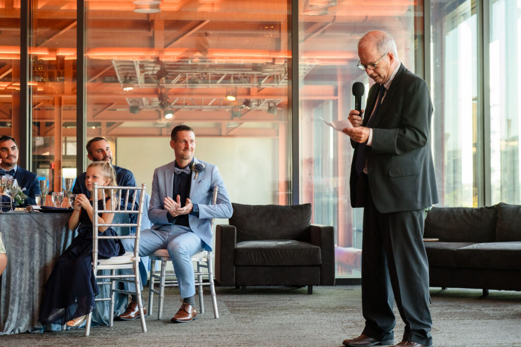 An older man in a suit speaks into a microphone while standing. Guests seated at a table, including a young girl and a man in a light blue suit, watch and smile. The room has large glass windows and modern decor.