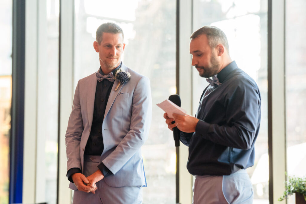 Two men in suits stand indoors; one reads from a paper while holding a microphone, and the other listens attentively. Large windows with an urban backdrop are behind them.