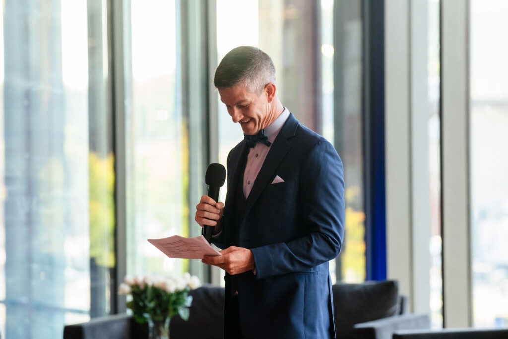 A man in a suit and bowtie stands indoors, holding a microphone and reading from a piece of paper, smiling as he prepares to speak.
