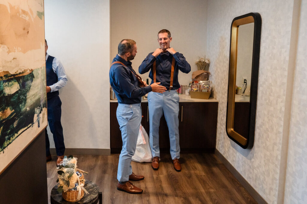 Two men in dress shirts and suspenders stand in a room, adjusting their outfits and smiling. A third person is partially visible on the left. The room has a mirror, counter, and artwork on the wall.