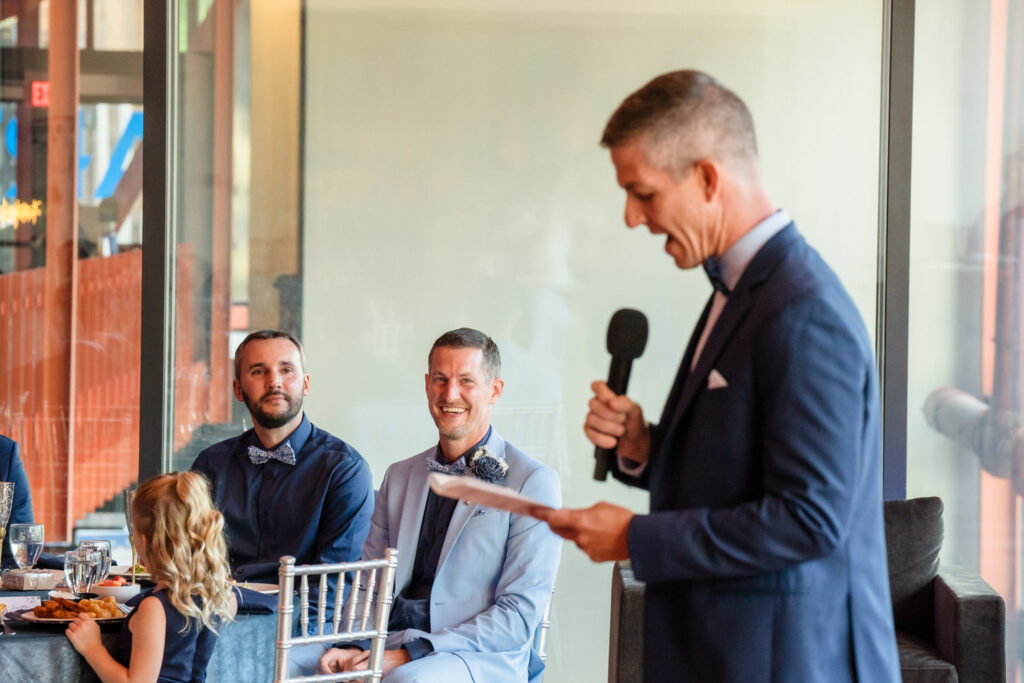 A man in a suit gives a speech with a microphone, while two seated men and a young girl listen and smile at a formal indoor event.