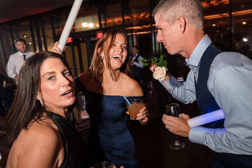 Three adults in formal attire dance at a lively indoor event. One woman holds a drink and smiles, the man has a rose and glow stick, and another woman looks toward the camera. Warm lighting and people appear in the background.