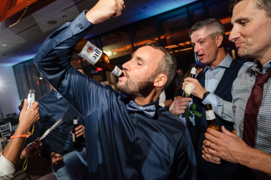 A man in a blue shirt and bow tie drinks from a beer bottle at a lively indoor party, surrounded by other men also holding beers.