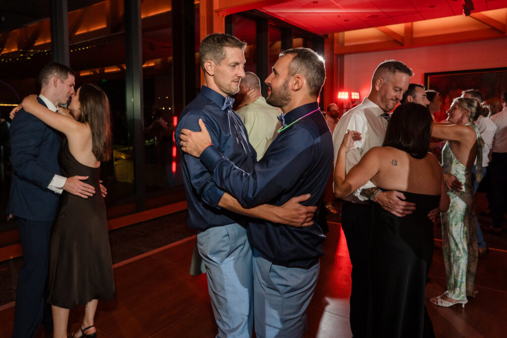 Two men dance together in the center of a warmly lit room, surrounded by other couples also slow dancing, creating a joyful, celebratory atmosphere.