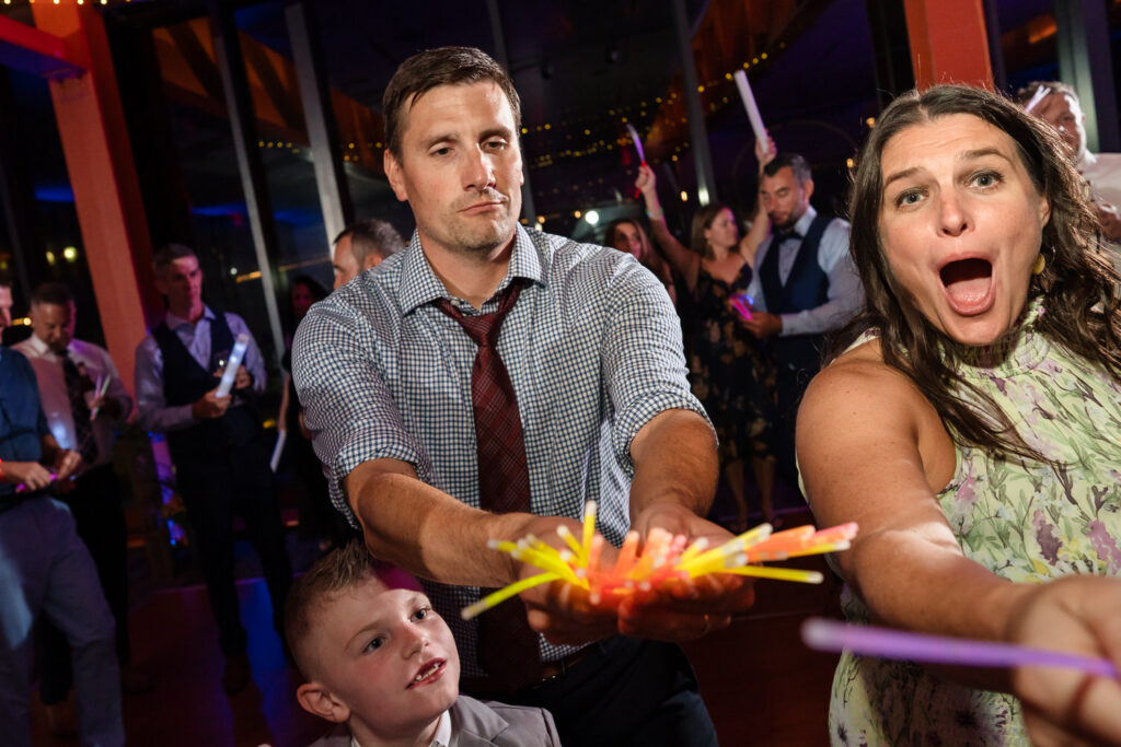 A man and woman excitedly hold glowing sticks at a lively indoor party, with a young boy in front. People are dancing and having fun in the colorful, festive background.