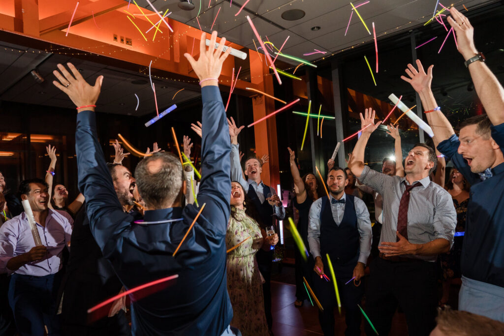 A group of people joyfully dance and throw colorful glow sticks in the air at an indoor party, with their hands raised and big smiles on their faces.
