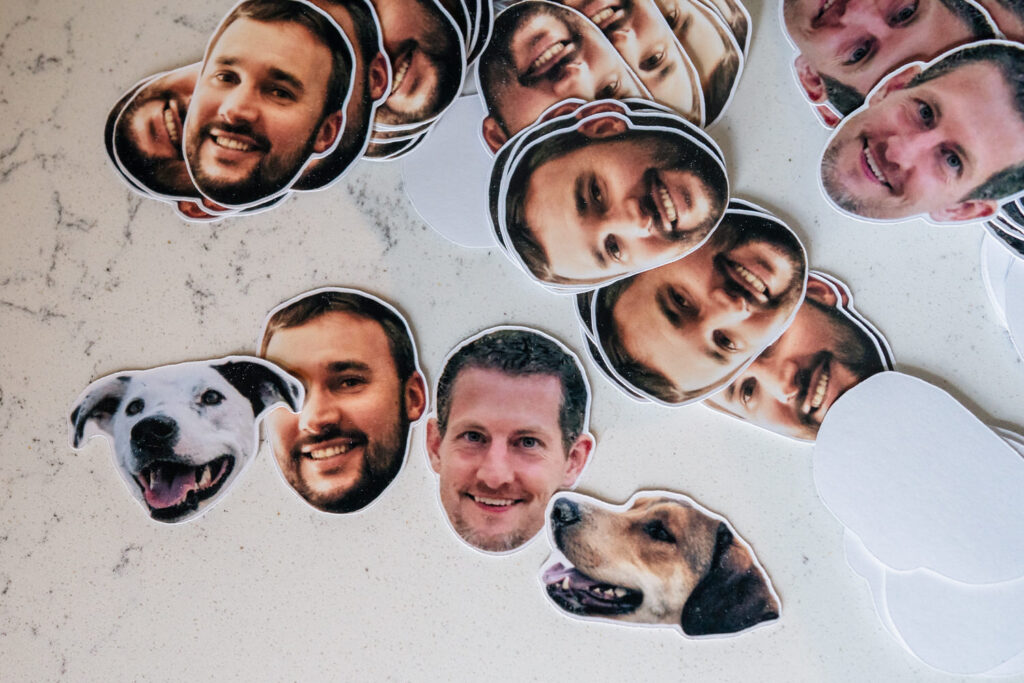 Several cutouts of human faces and two dog faces are scattered on a light-colored surface. The cutouts are made of paper and appear to be printed photo stickers.