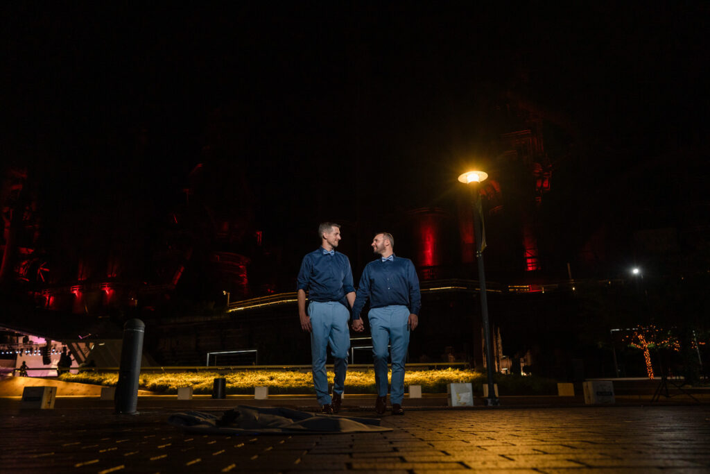 Two men holding hands and smiling at each other while walking outside at night under a streetlamp, with soft yellow and red lighting illuminating the background.