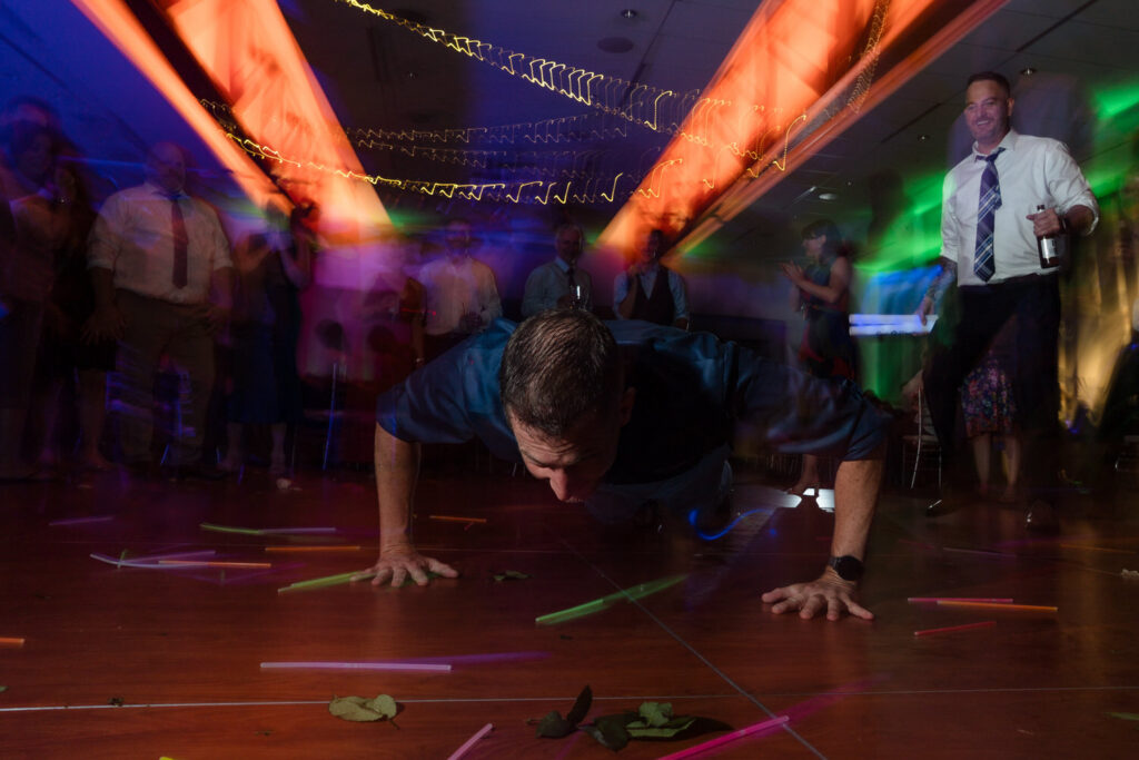 A man does push-ups on a dance floor with colorful lights and blurred party guests around him. Glow sticks and leaves are scattered on the floor.