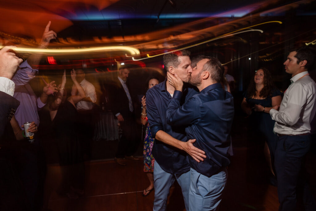 Two men kiss and embrace on a dance floor at a lively party, surrounded by people who are dancing and celebrating, with colorful light trails in the background.