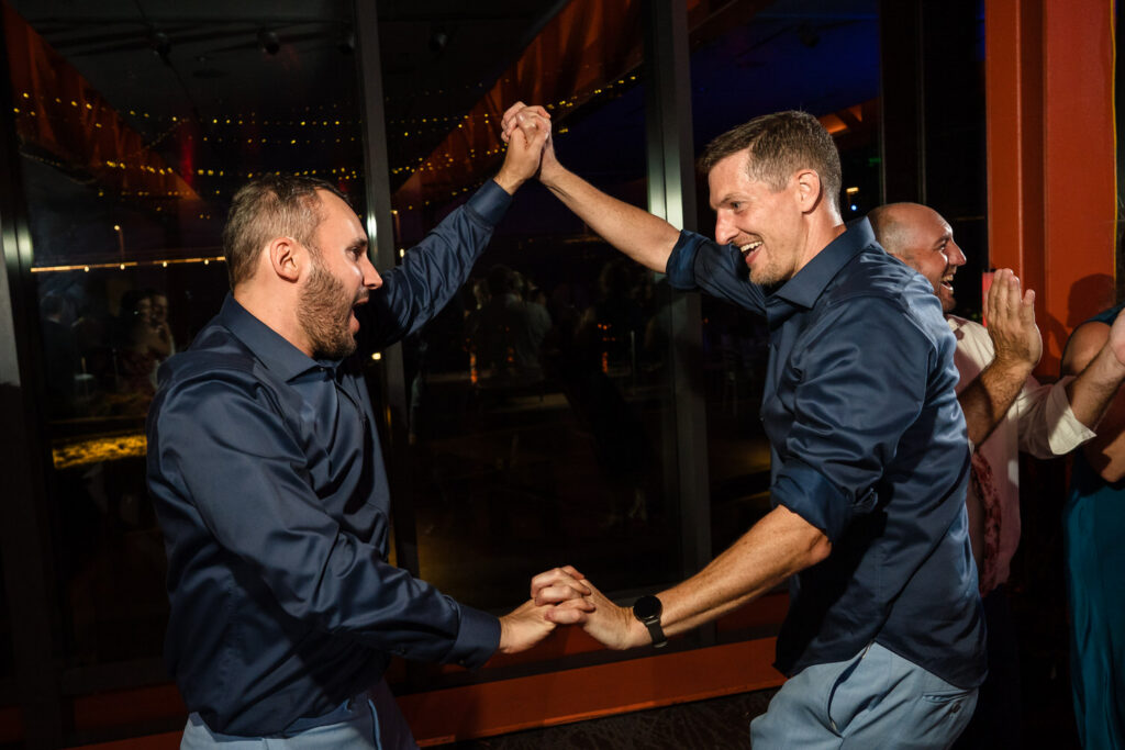 Two men in matching navy shirts are smiling and dancing together at a lively indoor event, holding hands and raising their arms. Other people are clapping and watching in the background.