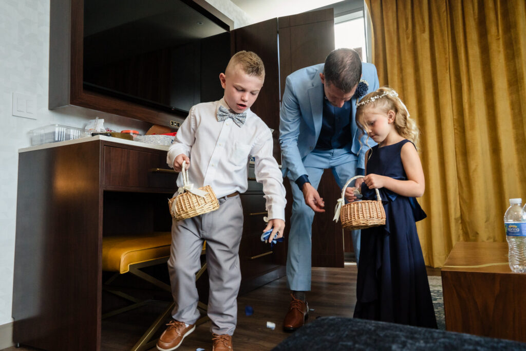 A boy and girl in formal attire hold baskets, while a man in a blue suit bends down to pick up items from the floor in a room with yellow curtains.