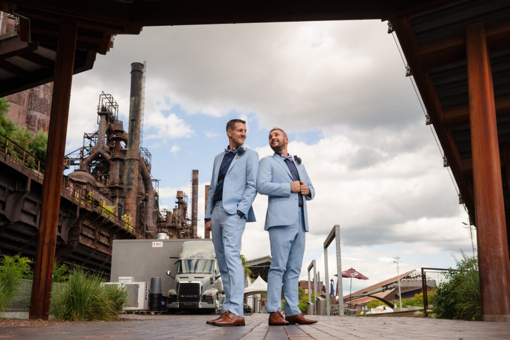 Two men in light blue suits stand back to back, smiling, at an industrial site with metal structures and cloudy sky in the background.