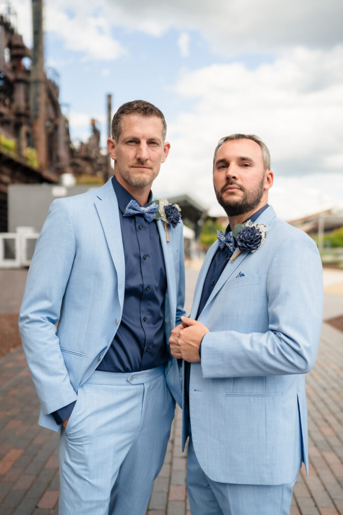 Two men in light blue suits with navy shirts and bow ties stand outdoors on a brick path. Both wear boutonnières and look at the camera. Industrial buildings and a cloudy sky are visible in the background.