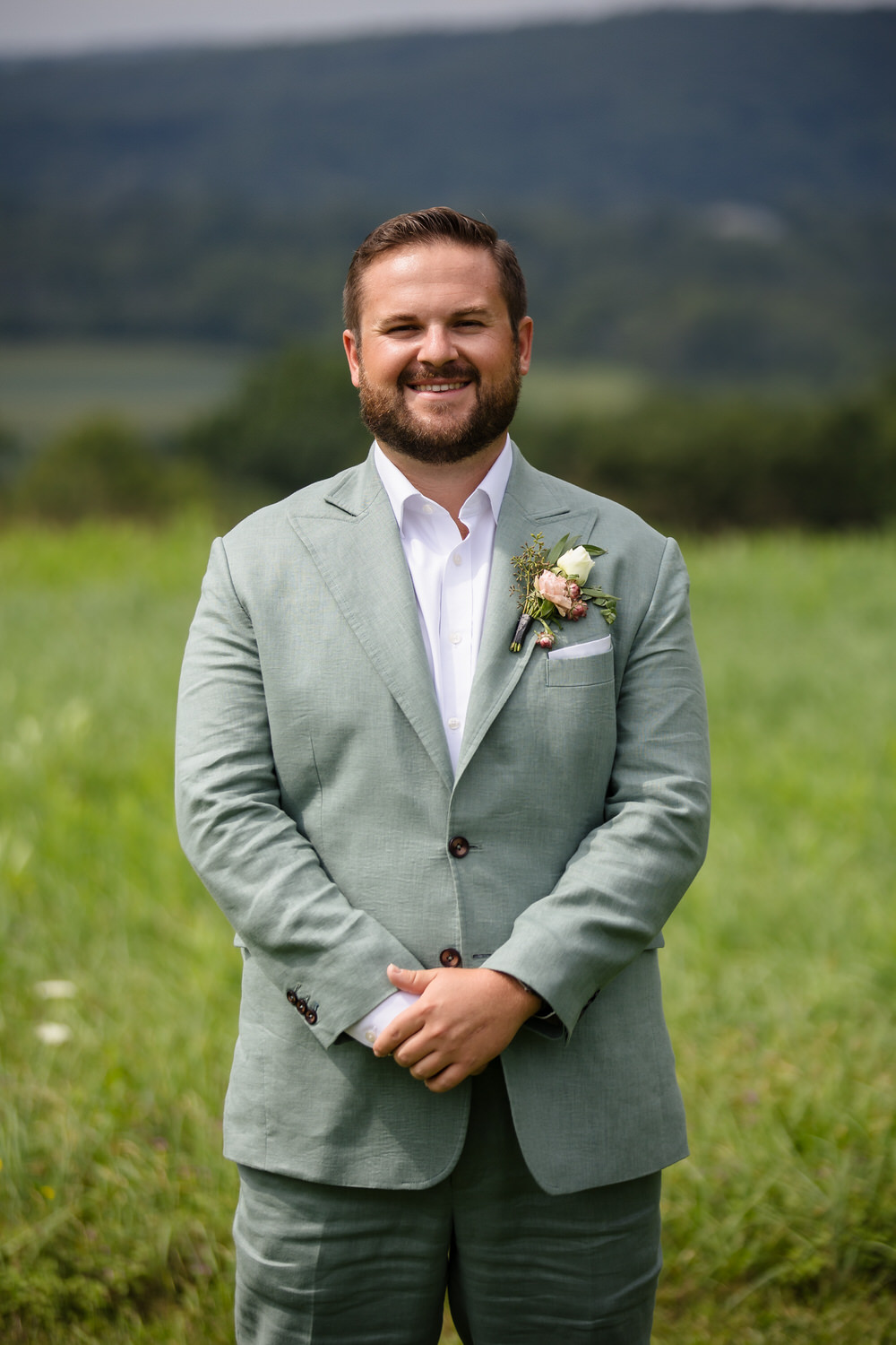 A man in a light green suit and white shirt stands smiling outdoors in a grassy field, with hills and trees in the background. He wears a floral boutonniere on his jacket.