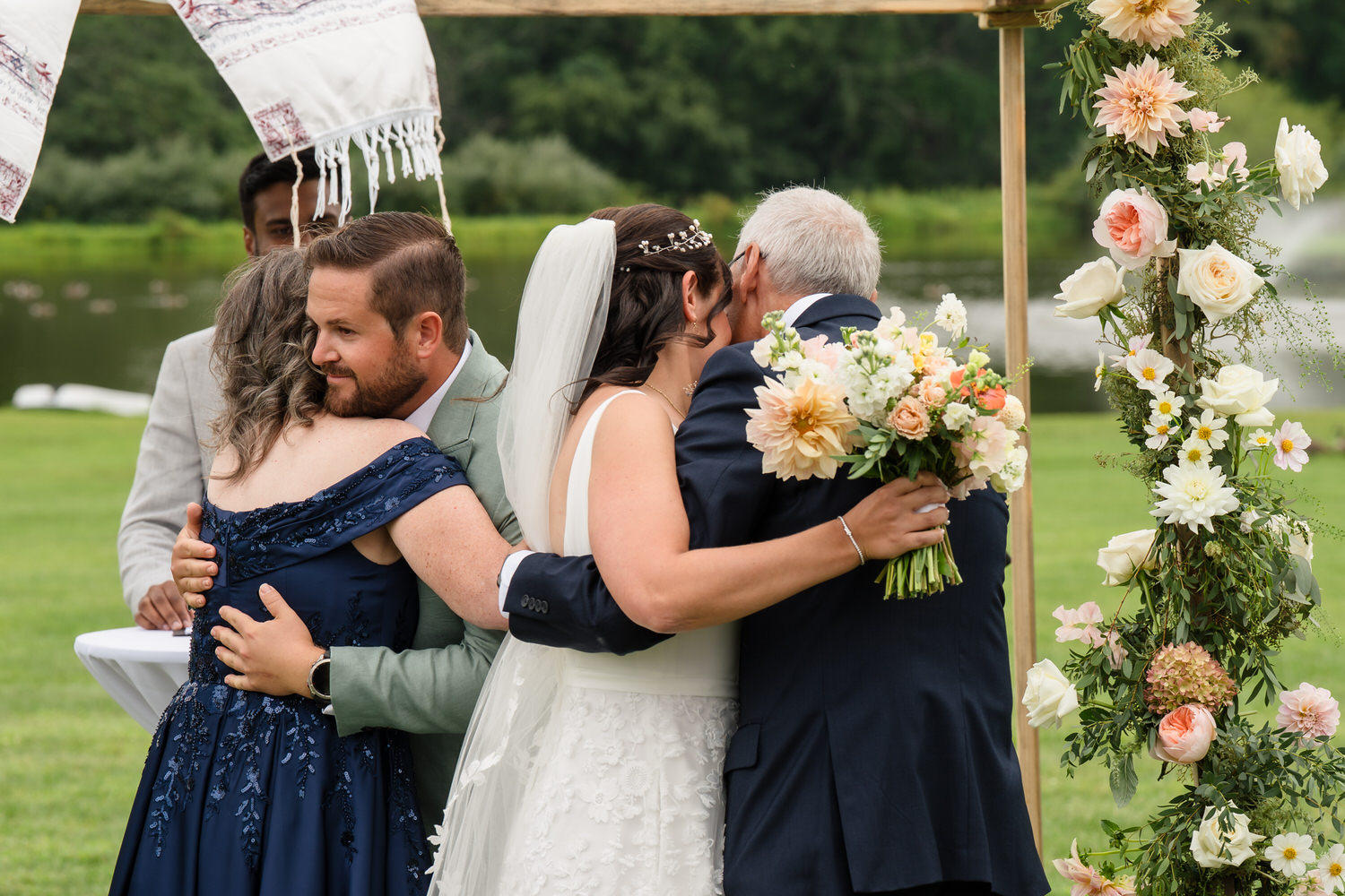 Two couples embrace during an outdoor wedding ceremony, with floral decorations and greenery in the background. The bride holds a bouquet of flowers while hugging an older man.