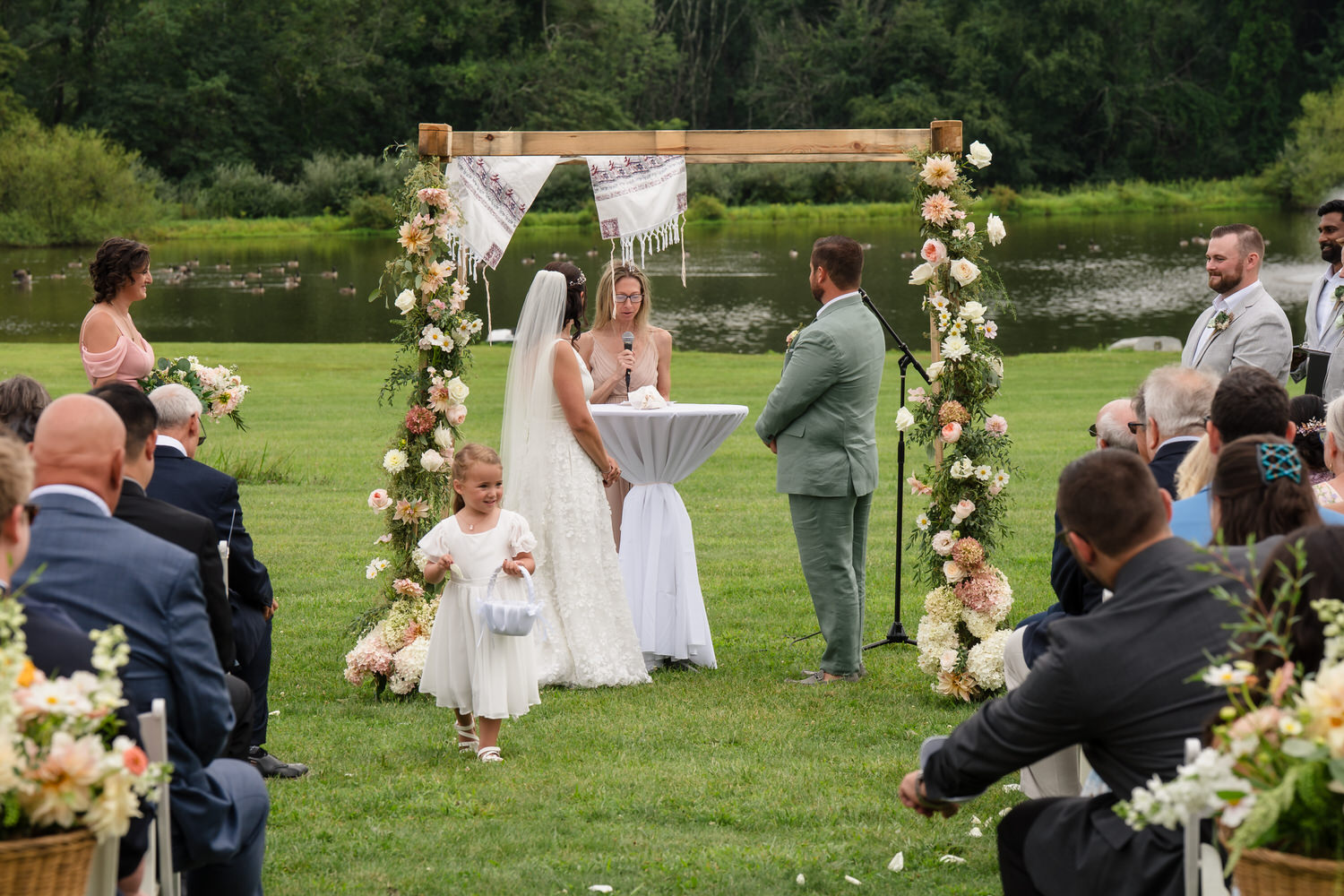A bride and groom stand under a floral arch by a lake, facing an officiant. Guests are seated outdoors, and a young flower girl in white walks down the aisle, holding a basket.
