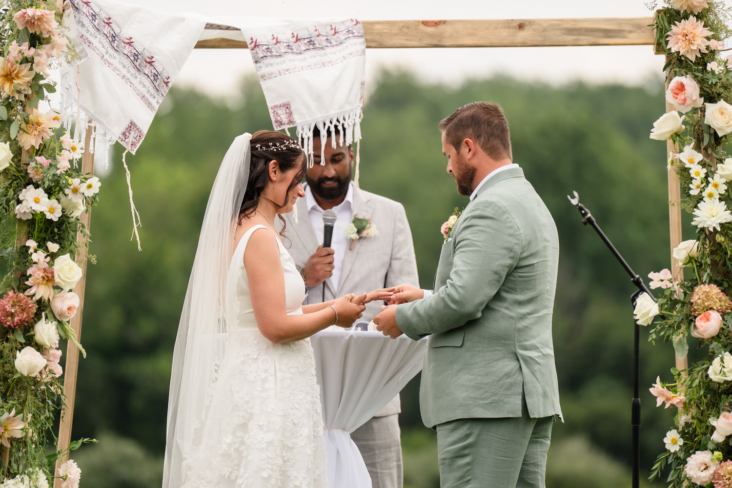 A bride and groom exchange rings at an outdoor wedding ceremony, standing under a floral arch, with an officiant in the background holding a microphone.